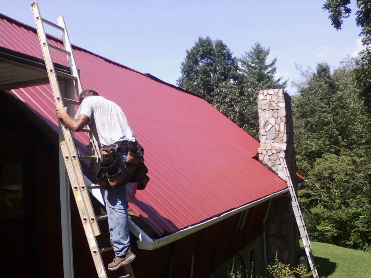 Roofer on a ladder beside a red metal roof and stone chimney — Soffit & Fascia Repair by Roofing Brothers in Harper Woods